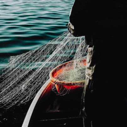 Vi ser en fiskare som står i en roddbåt och drar upp ett fiskenät av plast från vattnet