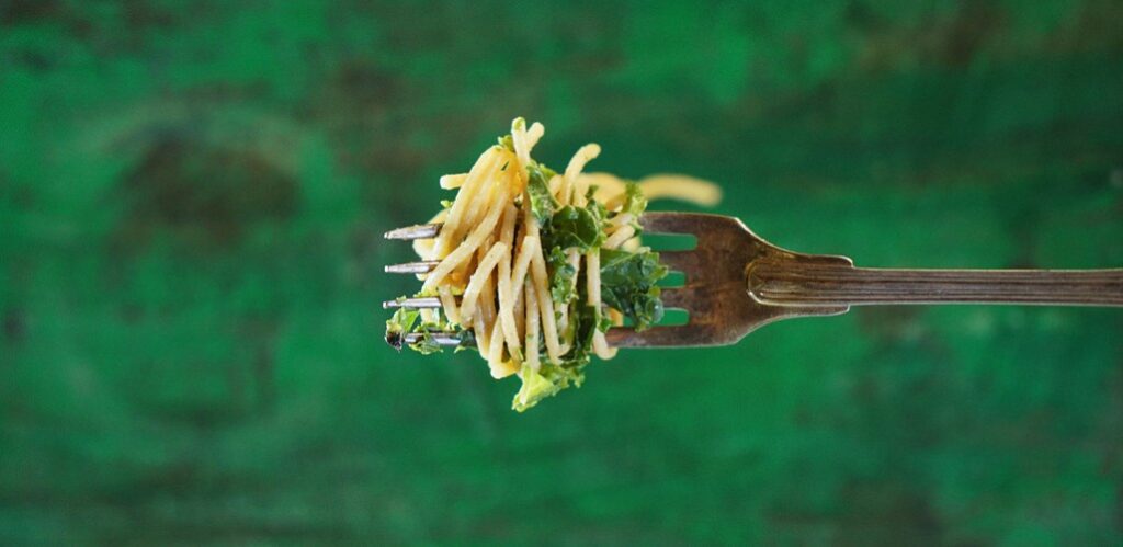 Närbild på en gaffel med spaghettis och grönkål. Grön bakgrund.